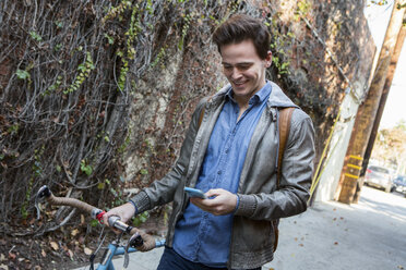 Young man strolling with cycle on sidewalk reading smartphone texts - ISF06963