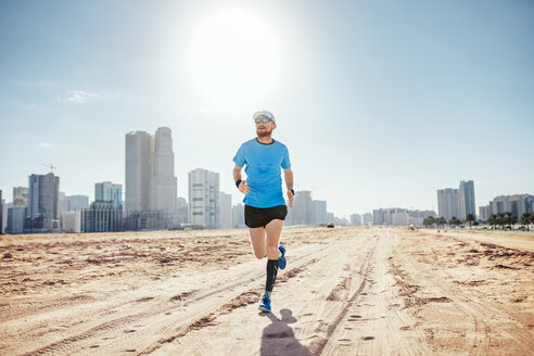 Vorderansicht eines erwachsenen Mannes in voller Länge, der auf Sand neben Wolkenkratzern läuft, Dubai, Vereinigte Arabische Emirate - ISF06969