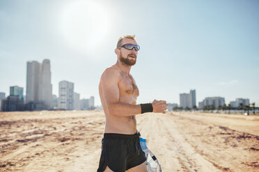 Mann mit nacktem Oberkörper und Sonnenbrille im Sand vor Wolkenkratzern, Dubai, Vereinigte Arabische Emirate - ISF06970