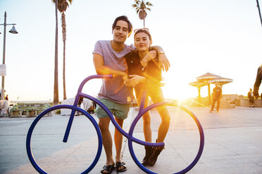 Portrait of young couple leaning against bicycle sculpture at coast, Venice Beach, California, USA - ISF07002