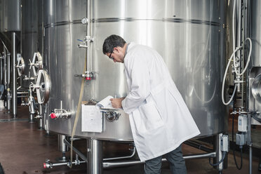 Side view of brewer in brewery wearing lab coat writing on clipboard - ISF07006