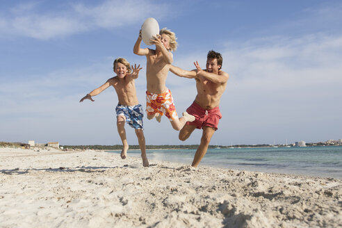 Junge, der mit einem Rugbyball springt und von seinem Bruder und seinem Vater am Strand verfolgt wird, Mallorca, Spanien - CUF18543