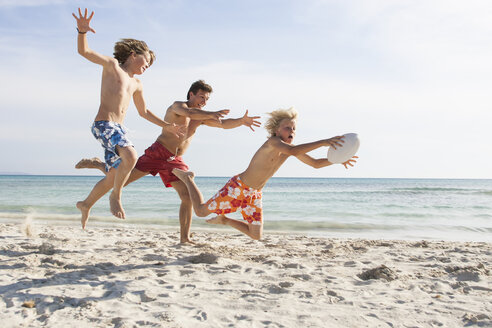 Junge und Vater jagen Bruder mit Rugbyball am Strand, Mallorca, Spanien - CUF18544