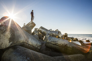 Junger Mann mit Skateboard auf dem Wellenbrecher stehend, Blick zum Meer - ISF07115