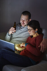 Mid adult couple sitting on sofa in evening eating popcorn and looking at laptop - ISF07117