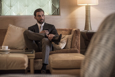 Young businessman sitting on hotel lobby sofa using smartphone - CUF18943