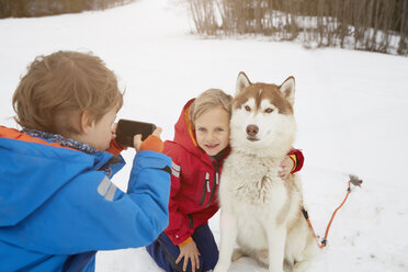 Boy taking smartphone photo of brother and husky in snow, Elmau, Bavaria, Germany - CUF18968