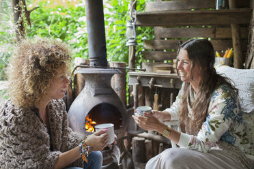 Two female friends drinking coffee in open cabin with wood stove - ISF07406