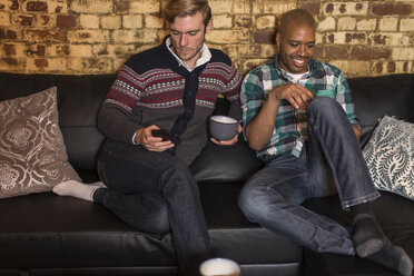 Male couple sitting on sofa, looking at smartphones - ISF07421