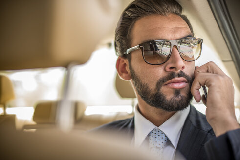 Junger Geschäftsmann mit Sonnenbrille, der auf der Rückbank eines Autos mit seinem Smartphone spricht, Dubai, Vereinigte Arabische Emirate - CUF19115