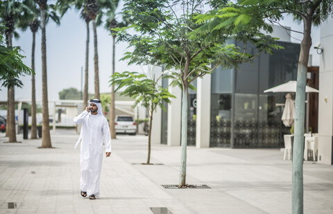 Mann in traditioneller Kleidung aus dem Nahen Osten geht auf der Straße und spricht mit seinem Smartphone, Dubai, Vereinigte Arabische Emirate, lizenzfreies Stockfoto