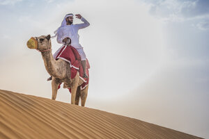 Mann in traditioneller nahöstlicher Kleidung reitet auf einem Kamel in der Wüste, Dubai, Vereinigte Arabische Emirate - CUF19149