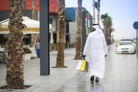 Rückansicht eines Mannes mit Dishdasha, der mit Einkaufstüten die Straße entlanggeht, Dubai, Vereinigte Arabische Emirate, lizenzfreies Stockfoto