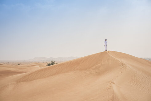 Junger Mann aus dem Nahen Osten in traditioneller Kleidung, der von einer Wüstendüne aus nach draußen schaut, Dubai, Vereinigte Arabische Emirate - CUF19212