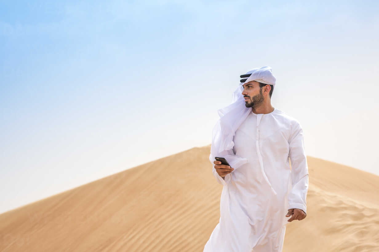 Middle eastern man wearing traditional clothes using smartphone on desert  dune, Dubai, United Arab Emirates stock photo