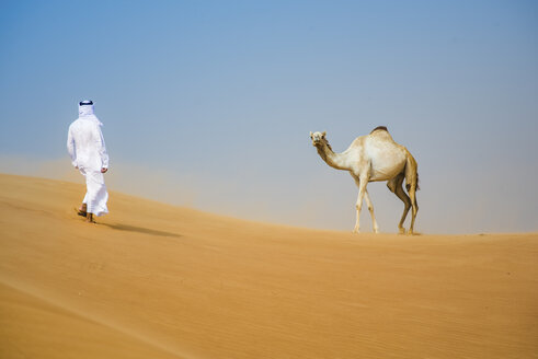 Mann aus dem Nahen Osten in traditioneller Kleidung geht auf ein Kamel in der Wüste zu, Dubai, Vereinigte Arabische Emirate - CUF19218