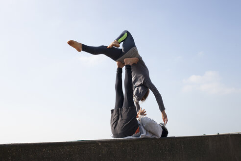 Man and woman practicing acrobatic yoga on wall against blue sky - CUF19328