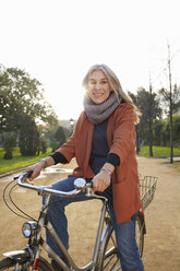 Mature woman in park on bicycle looking at camera smiling - CUF19365