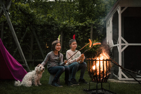 Zwei Mädchen mit Federkopfschmuck, grillen Würstchen am Lagerfeuer, Hund sitzt auf der Wiese - MOEF01216