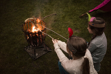 Zwei Mädchen mit Federkopfschmuck, die über einem Lagerfeuer Würstchen grillen - MOEF01217