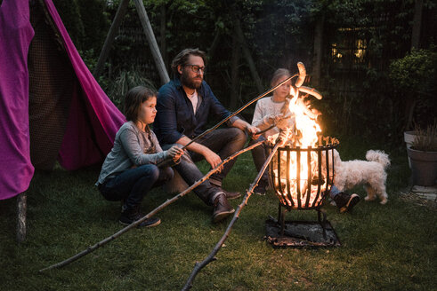 Man and two girls grilling sausage over camp fire in garden - MOEF01218