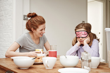 Mid adult woman at breakfast table with daughter wearing sleep mask - CUF19528