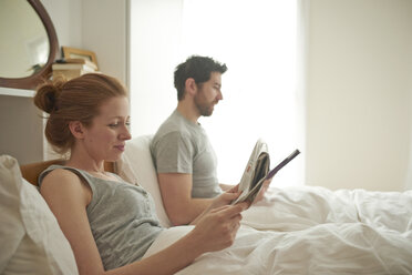 Mid adult couple reading digital tablet and broadsheet in bed - CUF19562