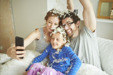 Eine verspielte Familie fängt einen lustigen Moment im Bett ein, bedeckt mit Federn von einer Kissenschlacht, während sie ein Selfie macht, lizenzfreies Stockfoto
