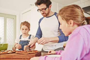 Mid adult man slicing pizza for daughters at kitchen bench - CUF19578