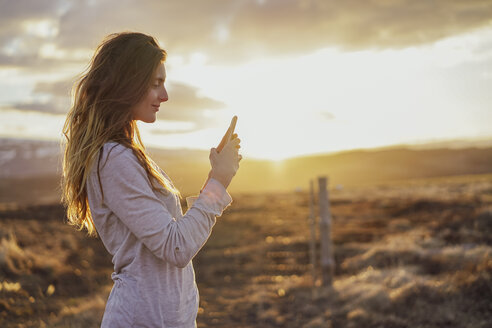 Island, junge Frau mit Smartphone bei Sonnenuntergang - AFVF00555