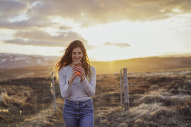 Island, junge Frau mit Smartphone bei Sonnenuntergang - AFVF00556