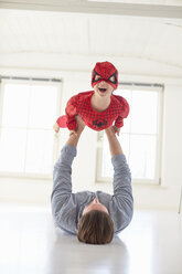 Father lying on floor holding up son wearing superhero costume - CUF19608