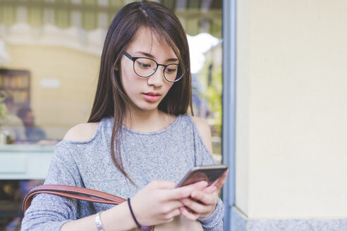 Junge Frau, die vor einem Straßencafé eine SMS auf ihrem Smartphone schreibt - CUF19853