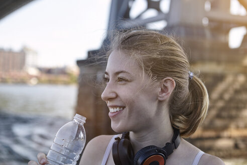 Junge Frau am Fluss, mit Kopfhörern um den Hals, hält eine Wasserflasche, New York City, USA - CUF19883