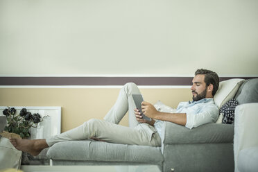 Junger Mann auf einer Chaiselongue in einem Hotelzimmer mit digitalem Tablet, Dubai, Vereinigte Arabische Emirate - CUF19987