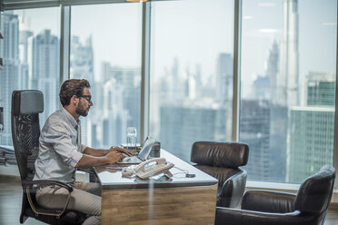 Businessman using laptop at desk with window view of city, Dubai, United Arab Emirates - CUF19989