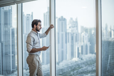Businessman with smartphone staring through window with skyscraper view, Dubai, United Arab Emirates - CUF19990