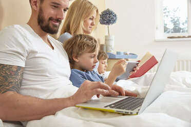 Mother and father in bed with sons using technology, reading book - CUF20027