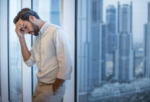Geschäftsmann mit Hand auf der Stirn am Fenster mit Blick auf einen Wolkenkratzer, Dubai, Vereinigte Arabische Emirate - CUF20058