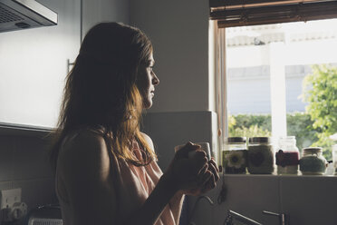 Junge Frau macht eine Kaffeepause und blickt durch das Küchenfenster - CUF20074