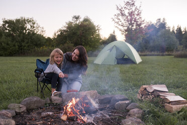 Mother and daughter sitting beside campfire, cooking sausage over fire - ISF07470