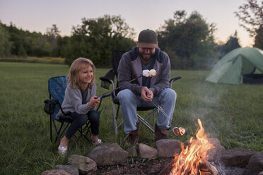 Father and daughter sitting beside campfire, toasting marshmallows over fire - ISF07473