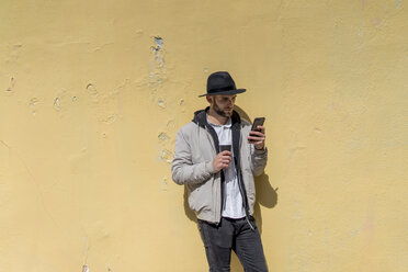 Bärtiger Mann mit schwarzem Hut, an die Wand gelehnt, mit Smartphone, Kaffee zum Mitnehmen - AFVF00588