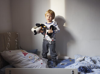 Portrait of little boy standing on bed with his soft toys - MUF01531