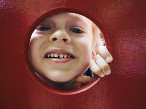 Porträt eines lächelnden kleinen Jungen, der durch ein Loch auf einem Spielplatz schaut - MUF01540