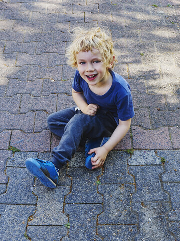 Porträt eines lächelnden, blau gekleideten kleinen Jungen auf dem Bürgersteig sitzend, lizenzfreies Stockfoto