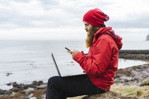 Island, Nordisland, Mann mit Laptop sitzt am Meer und telefoniert - AFVF00605