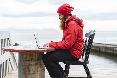 Island, Nordisland, Mann sitzt vor dem Meer und benutzt einen Laptop, lizenzfreies Stockfoto