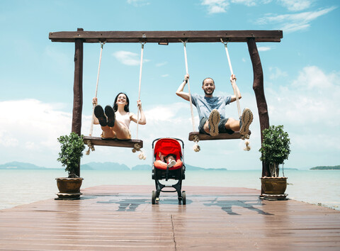 Thailand, Koh Lanta, happy parents on wooden swings in front of the sea and sleeping baby in a stroller stock photo