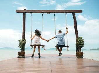 Thailand, Koh Lanta, back view of couple sitting on swings in front of the sea holding hands - GEMF02048